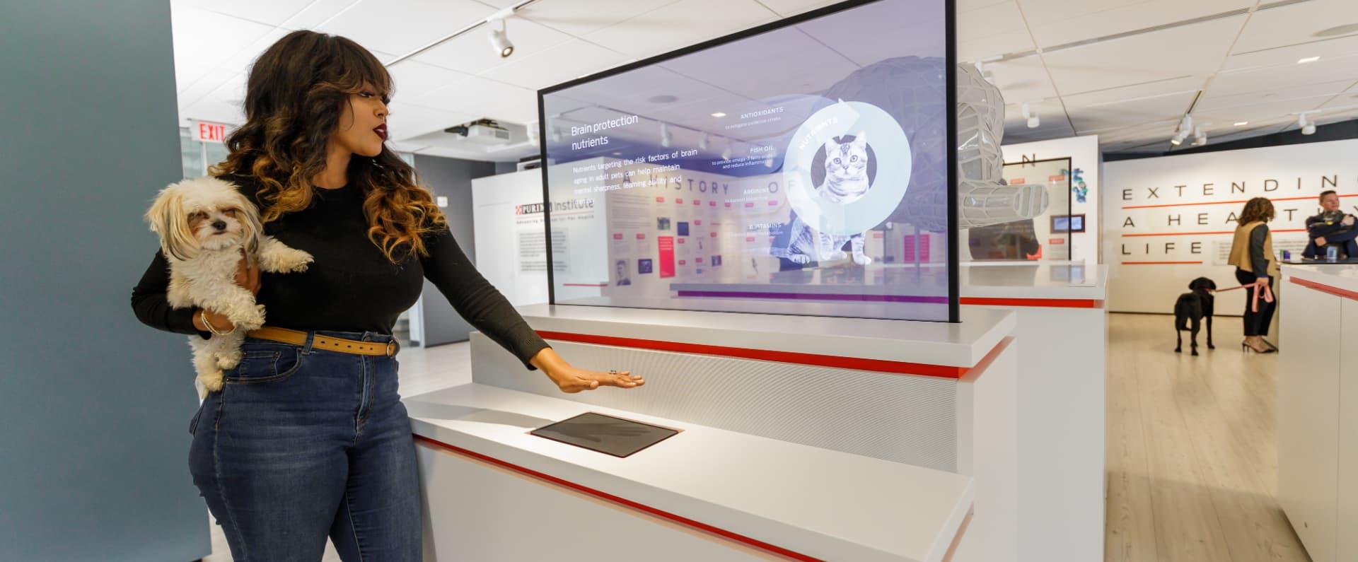 Black woman with brown hair holding small dog clicking on interactive monitor.